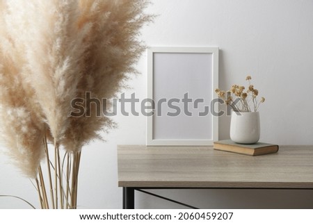 White frame mockup on table with books, ceramic vase with yellow dry Everlasting flowers. White wall background.