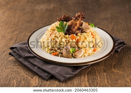 Pilaf with chicken drumstick in a light plate on a napkin with garlic and fresh herbs on a dark wooden background. A traditional oriental dish. Side view with copy space. Horizontal orientation. Royalty-Free Stock Photo #2060463305