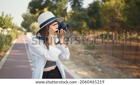 Young woman photographer wanderer. Happy young woman using modern digital camera for taking photos while walking. Female tourist in summer hat and clothes saving memories from trip