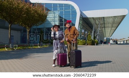 Elderly old husband and wife retirees tourists near airport terminal get video call use mobile cell phone, posing for social media selfie photos. Mature grandmother grandfather with luggage suitcases