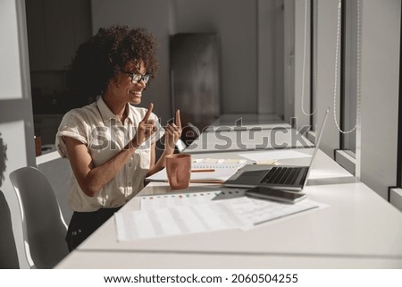 Happy pretty lady learning and communicating in sign language online while sitting in the office Royalty-Free Stock Photo #2060504255