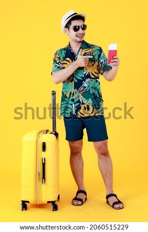 Enjoyed young Asian man in colorful Hawaiian shirt holds and point finger to  passport near suitcase. Full body studio portrait on yellow background. Happy summer vacation travel concept