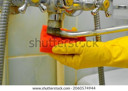 Side view of woman's hand in rubber glove with sponge wiping rusty, old chrome faucet. Close-up housewife cleaning plumbing fixtures from dirt in bathtub indoors. Household cleaning, bathroom washing. Royalty-Free Stock Photo #2060574944