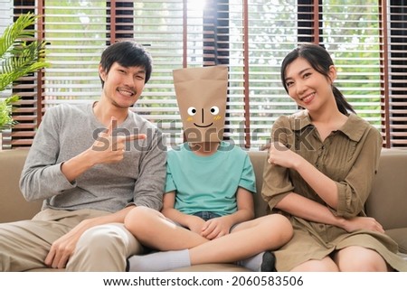 concept of family face emotions,funny humor dad mom hand pointing to his son wearing paper bag with smiling happiness face drawing on sofa,family siting together on sofa in living room home background