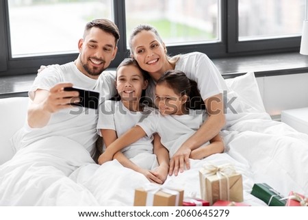 family, winter holidays and people concept - happy mother, father and two daughters with christmas gifts taking selfie on smartphone in bed at home