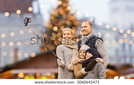 winter holidays, technology and people concept - happy family taking picture with smartphone and selfie stick over christmas market background