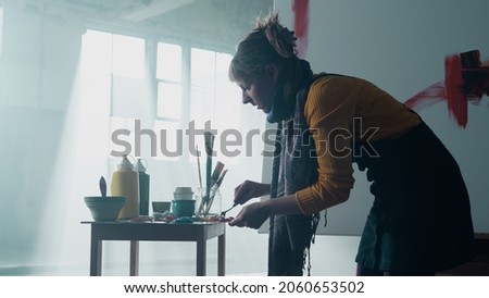 Isolated Beautiful Caucasian Girl, Mixing Colours on Her Palette, Standing Alone in an Industrial Warehouse With Big Windows, Sun Shining Through Them. Painting an Abstract Painting, Wearing Overalls.