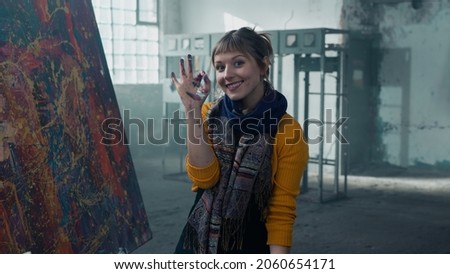 Isolated Beautiful Caucasian Female Painter Standing Next to her Masterpiece Contemporary Abstract Painting, Gesturing an Okay Sign With Her Hand, Looking Directly at the Camera. Warehouse Background.