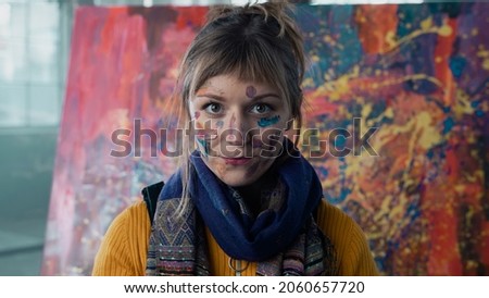 Beautiful Caucasian Painter Looking Directly at the Camera,  Standing in an Industrial Building With Her Face Covered in Colourful Paint, Wearing a Scarf Around Her Neck, Abstract Painting Behind Her.