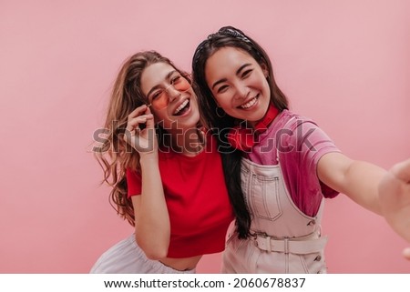 Close-up shot of two interracial young women taking vivid selfie against pink background. Brunette asian with headphones and blonde ukrainian in sunglasses are happy together. 