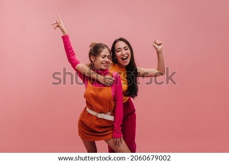 Smiling asian young woman hugs caucasian girlfriend joyfully looking into camera. Shot of two girls fooling around in photo studio on pinkish background. Holiday life concept Royalty-Free Stock Photo #2060679002