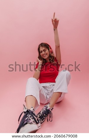 Young European cute girl sitting on floor touching camera with her shoes on pink background. Blonde with natural makeup in bright crop top is drowning in music and posing for cozy studio.