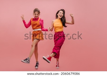 Full-length young asian and ukrainian girl are dancing amusingly on pink background with space for text. Wonderful girlfriends have fun holding hands, dressed in orange and crimson clothes.