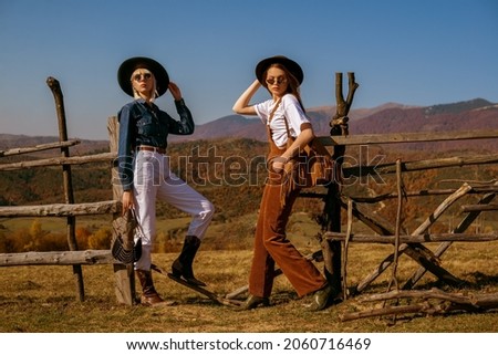 Two fashionable models wearing stylish autumn outfits with trendy hats, sunglasses, bags, cowboy boots, posing in mountains. Full length outdoor portrait. Copy, empty space for text