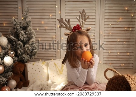 Cute girl with blonde hair in a New Year's costume on the background of a Christmas tree eating an orange