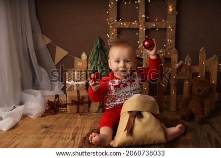 A cute kid in a New Year's costume on the background of a Christmas tree and garlands opens a New Year's gift and takes out red Christmas balls