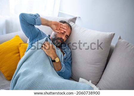 Cropped shot of a mature man suffering with flu while sitting wrapped in a blanket on the sofa at home. It's the season of sneezes. Man blowing his nose while lying sick in bed
