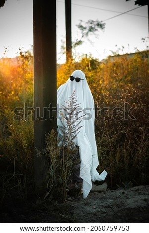 A man dressed as a ghost from a white sheet is hiding behind a post by the road. Ghost Challend 2021. Halloween Celebration. Spooky season.