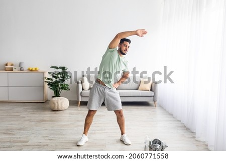 Full length of athletic young Arab man bending aside, doing exercises at home, copy space. Millennial Eastern man having domestic training, working out indoors during covid lockdown Royalty-Free Stock Photo #2060771558