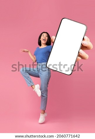 Check out this new mobile application. Excited young Asian lady jumping, shouting OMG, presenting smartphone with empty screen over pink studio background, mockup for website or app