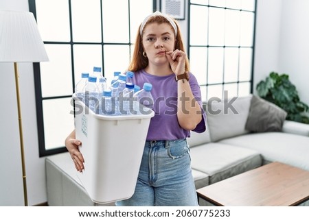 Young redhead woman holding recycling wastebasket with plastic bottles mouth and lips shut as zip with fingers. secret and silent, taboo talking 