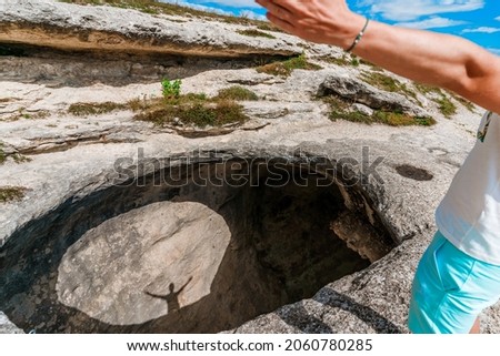 A man stands on the edge of a precipice in the form of a huge hole in the ground with his hands raised, casting a shadow into the hole. The concept of extreme travel and achievement of goals
