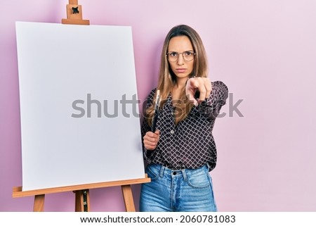 Beautiful hispanic woman standing by painter easel stand holding brushes pointing with finger to the camera and to you, confident gesture looking serious 