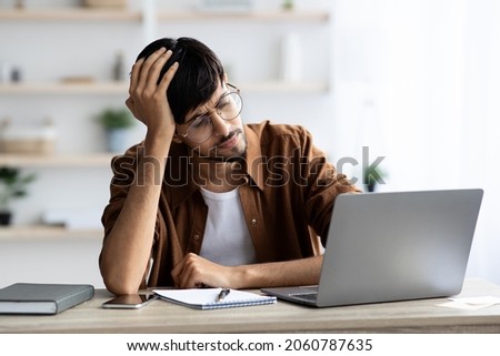 Exhausted indian man sitting in front of laptop at office, touching his head, suffering from burnout or financial crisis, young guy freelancer having difficulties with job, lack of clients, copy space Royalty-Free Stock Photo #2060787635