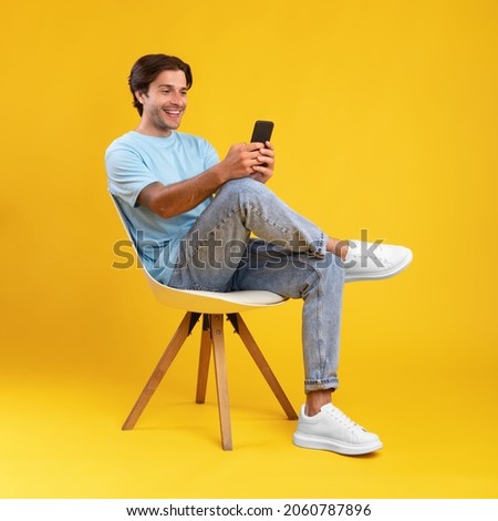 People And Technology Concept. Portrait of smiling young man using smartphone sitting on chair isolated on orange studio background. Excited casual guy chatting online, browsing social media Royalty-Free Stock Photo #2060787896