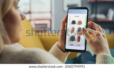 Beautiful Caucasian Female is Using Smartphone with Clothing Online Web Store to Choose and Buy Clothes from New Collection. Female Surfing the Net and Lying on Couch Sofa at Home Living Room.