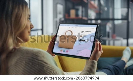 Beautiful Caucasian Female is Using Tablet Computer with Clothing Online Web Store to Choose and Buy Coat from New Collection. Female Surfing the Net and Lying on Couch Sofa at Home Living Room. Royalty-Free Stock Photo #2060794994