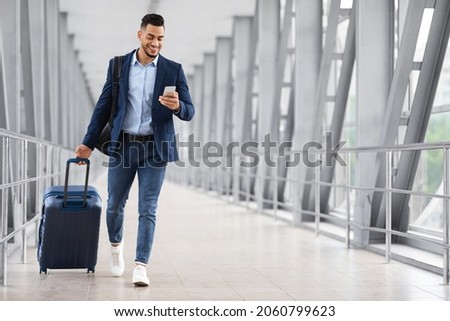 Online Check In. Handsome Arab Man Walking With Suitcase In Airport And Using Smartphone, Smiling Young Middle Eastern Guy Browsing Internet On Cellphone While Going To Flight Gate, Copy Space Royalty-Free Stock Photo #2060799623