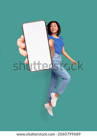 Full length of cheery Asian woman jumping and smiling, showing cellphone with empty space for mobile app or website on screen, turquoise studio background, mockup. Creative collage Royalty-Free Stock Photo #2060799689