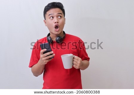 Portrait of shocked Asian man in red t-shirt looking away while holding mobile phone and a mug. Wow face expression. Isolated image on gray background