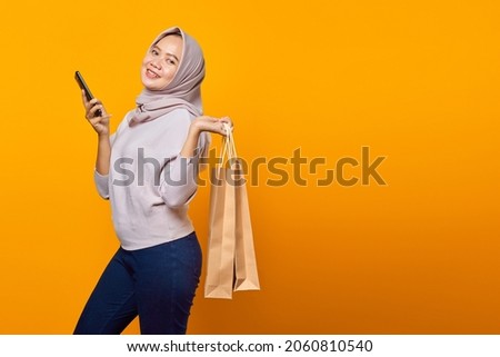 Portrait of cheerful asian woman holding phone and shopping bag over yellow background