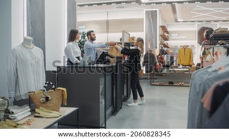 Clothing Store Checkout Cashier Counter: Beautiful Young Woman Buys Blouse from Friendly Retail Sales assistant, Receiving Bag with Purchases. Trendy Fashion Shop with of Designer Brands Royalty-Free Stock Photo #2060828345