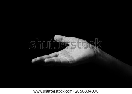 Black and white image of wrinkled male hand held out gesturing, with open palm up, isolated against a black background with dramatic lighting.