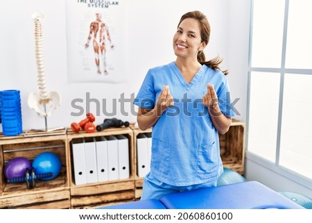 Middle age hispanic physiotherapist woman working at pain recovery clinic doing money gesture with hands, asking for salary payment, millionaire business 