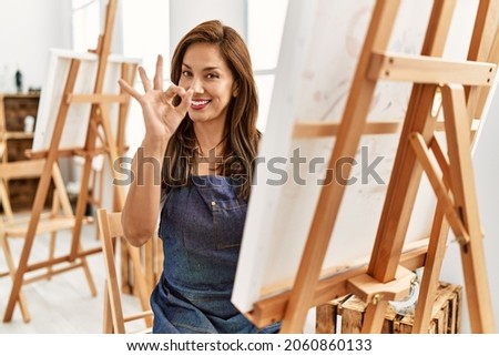 Young latin artist woman painting at art studio doing ok sign with fingers, smiling friendly gesturing excellent symbol 