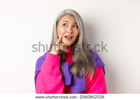 Close up of beautiful asian elderly woman in stylish clothes, making plans, looking aside and thinking, standing over white background