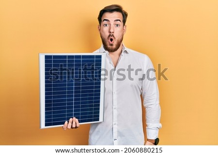 Handsome caucasian man with beard holding photovoltaic solar panel scared and amazed with open mouth for surprise, disbelief face 