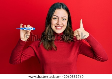 Young brunette woman holding thermometer smiling happy and positive, thumb up doing excellent and approval sign 