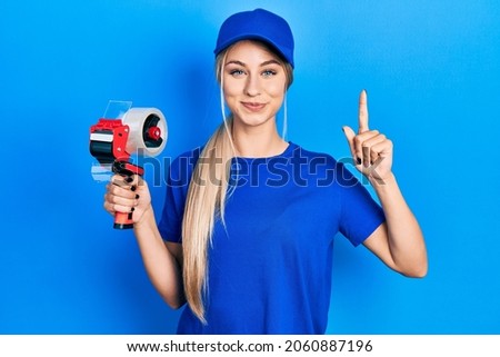 Young beautiful caucasian woman holding packing tape smiling with an idea or question pointing finger with happy face, number one 