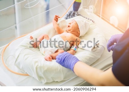 Unrecognizable nurse in blue gloves takes action and care for premature baby, selective focus on baby eye Newborn is placed in the incubator. Neonatal intensive care unit. Sun glare effect. Royalty-Free Stock Photo #2060906294