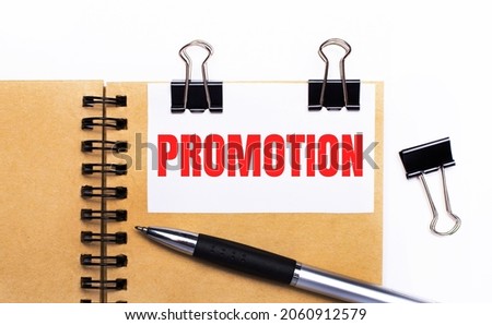 On a light background, a brown notebook with a pen, black clips and a white card with the text PROMOTION