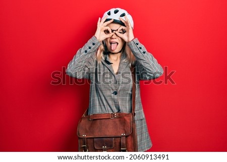 Beautiful caucasian blonde business woman wearing bike helmet doing ok gesture like binoculars sticking tongue out, eyes looking through fingers. crazy expression. 