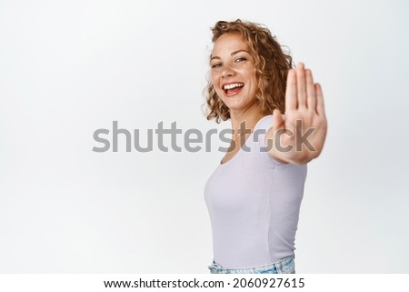 Smiling blond woman extend hand to shop stop sign, rejecting something with unbothered happy face expression, white background