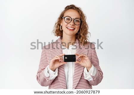 Smiling female entrepreneur in glasses showing credit card, standing in suit against white background