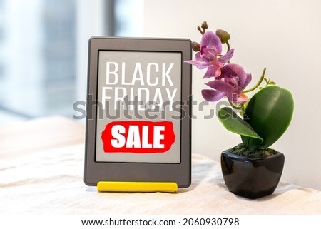 Black Friday Sale sign on e-reader electronic book next to artificial pink orchid on the kitchen table with a linen tablecloth by the window