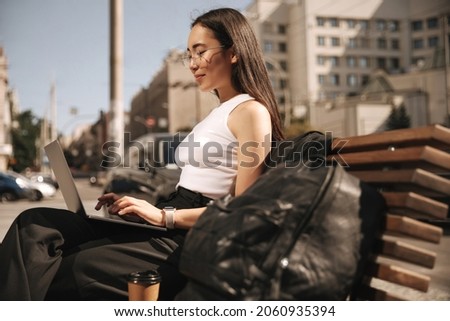 Close-up photograph of serious, focused young asian woman looking through information found on her laptop wearing glasses. Outdoors sits on bench. Freelance work, concept